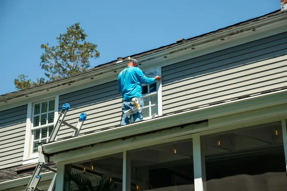 Painter on a ladder brushing fresh paint onto white wood trim of a New England colonial house